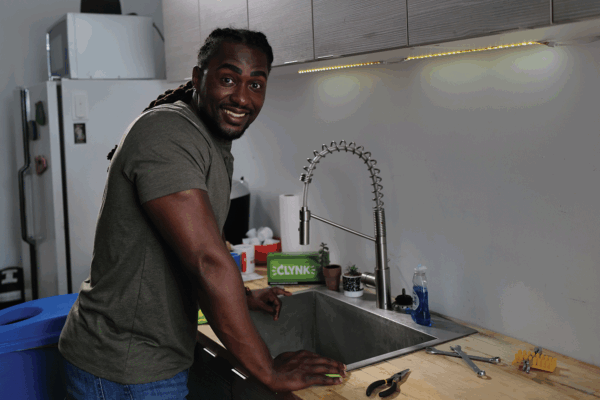 Man smiling while cleaning a kitchen sink with tools nearby on a wooden countertop.